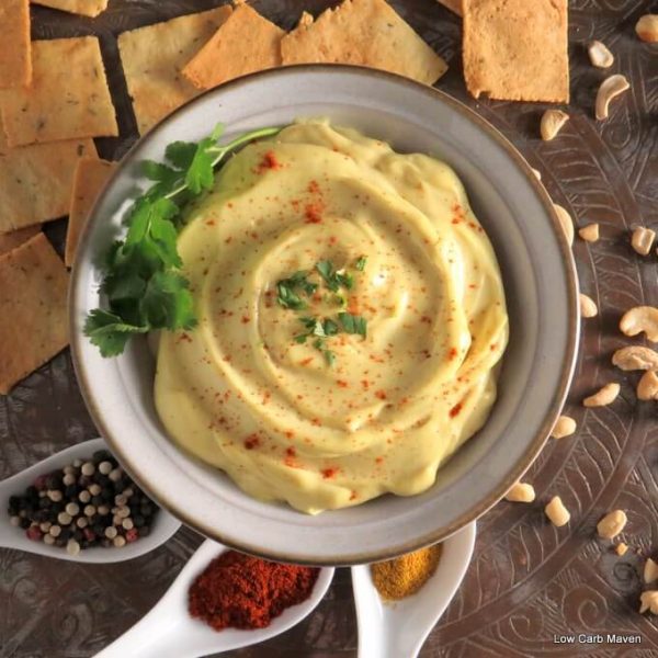 Creamy curry cashew cream dip in a bowl garnished with cilantro and chili powder with crackers, cashew nuts and white spoons of vibrant spices.