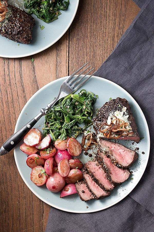 Sliced pan seared flat iron steak with a peppercorn crust and blue cheese butter on a light blue plate with pan fried radishes and sauteed radish greens.