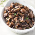 Quartered sauteed mushrooms in white wine sauce with thyme in a white dish.