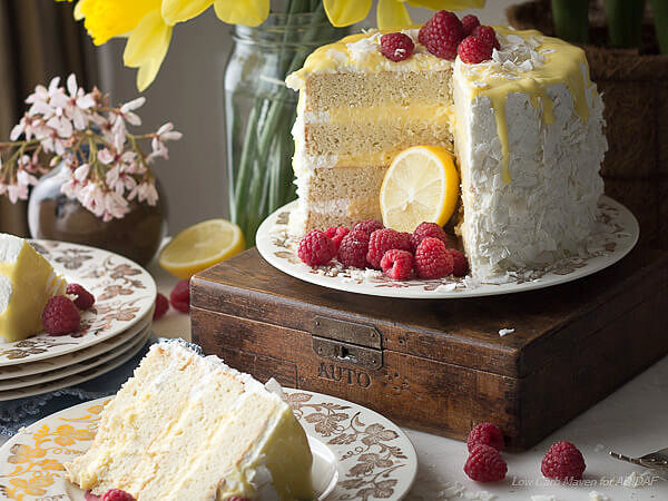 White layer cake filled with lemon curd, frosted with whipped cream and decorated with flaked coconut and raspberries sitting on a vintage china plate on an antique wodden box.