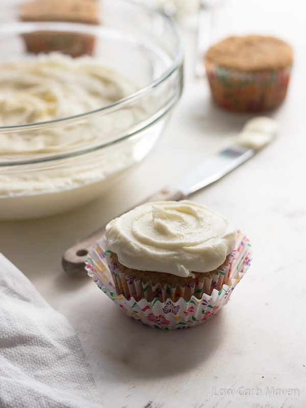 A cupcake in bright floral paper is topped with silky sugar free cream cheese frosting with a white towel to the left side, a small wooden handled stainless-steel spatula with frosting on it's tip is behind the cupcake as is a large clear glass bowl of cream cheese frosting and additional un-frosted cupcakes in the back ground.