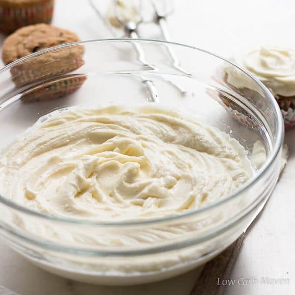 A large clear bowl of swirled sugar free cream cheese frosting with beaters placed behind and un-frosted cupcakes to the left of the beaters and a frosted cupcake to the right all on a white marble surface.