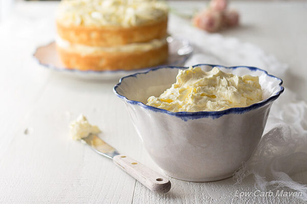 Low carb Sugar free French buttercream frosting in a white ruffled bowl with a blue edge between a small wooden handled spatula on the side with frosting on it's tip and a white piece of gauzy cheesecloth with a partially decorated cake in the background and pink roses.