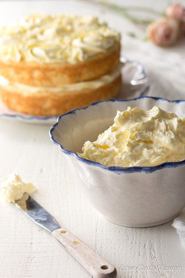Rich yellow sugar free French buttercream frosting in a white ruffled bowl with a blue edge and a small wooden handled spatula with frosting on it's tip to the left on a white wooden surface with a two layered naked cake behind with yellow buttercream rosettes on the top and pink roses in the background.