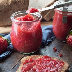 Jars of strawberry jam and toast on a wooded board with a blue napkin and a loaf of sliced bread behind.