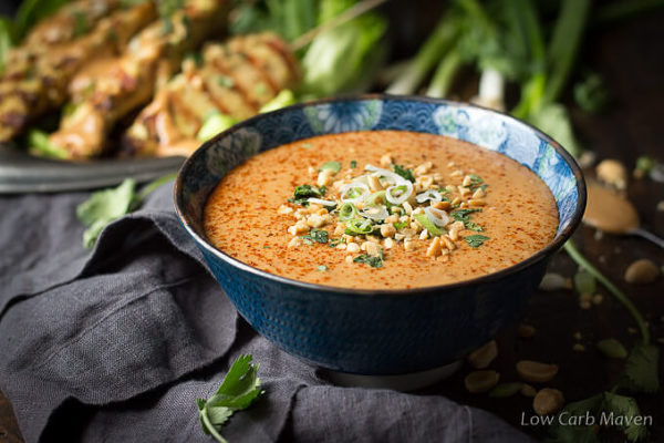 Peanut sauce garnished with cayenne pepper, cilantro, green onion and crushed peanuts in a blue Asian print bowl placed on a dark gray napkin with chicken satay on skewers, scallions and peanuts in the background.