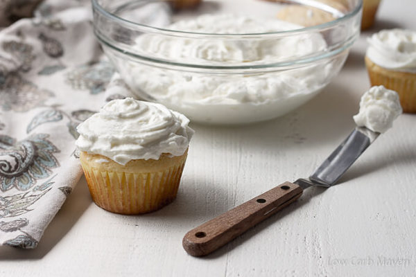 A cupcake slathered in whipped cream cheese frosting sits between a floral napkin and a small spatula with frosting with a glass bowl of cream cheese frosting and frosted cupcakes behind.