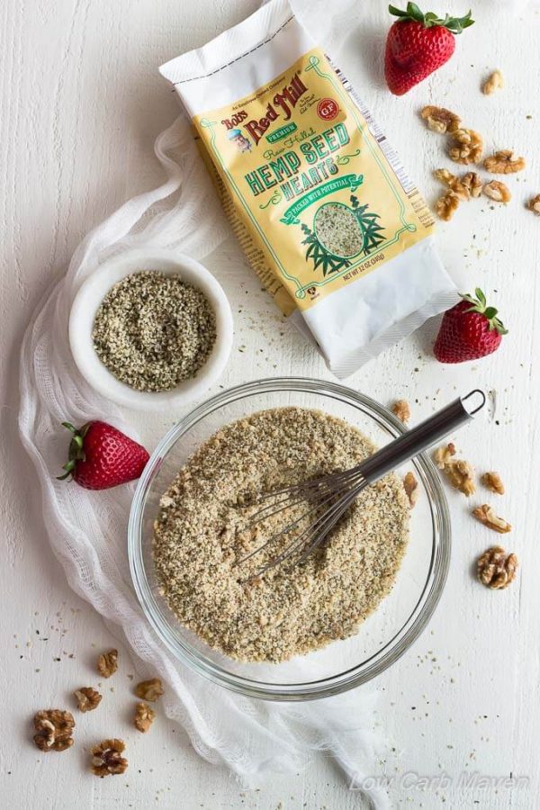 Ingredients for low carb hemp walnut pie or tart crust featuring ground hemp seeds and walnuts in a bowl, toasted walnuts on a marble surface with fresh strawberries and a package of Bob's Red Mill Hemp Seeds.