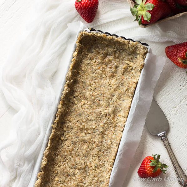 Hemp walnut crust pressed into a parchment lined rectangular tart pan with a sliver pie server to the right, a white cheesecloth to the left and fresh strawberries around.
