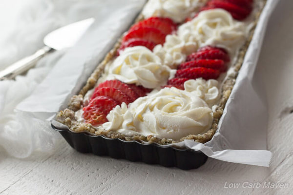A closeup of a strawberry dessert tart with cream cheese tart featuring a hemp and walnut crust in a black rectangular tart pan lined in white parchment decorated with sliced fresh strawberries and piped cream cheese roses.