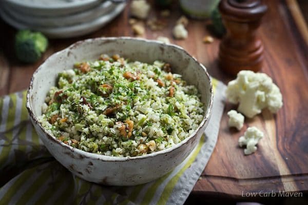 A low carb side of cauliflower and broccoli rice with walnuts in a bowl sitting on a wooden cutting board with a green striped napkin and a wooden pepper mill in the background.