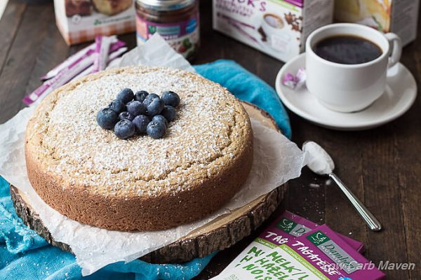 Sugar-free yellow cake topped with powdered sweetener and blueberries on a piece of parchment and round rustic cake stand with a blue napkin to the left and sugar free chocolate bars made with tagatesse to the right and sugar-free raspberry jam behind near a cup of coffee.