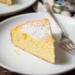 Slice of lemon ricotta cake with powdered sugar on a white plate with fork.