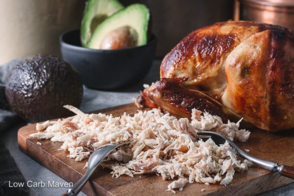 Shredded rotisserie chicken for chicken enchiladas on a cutting board with fork and avocados.