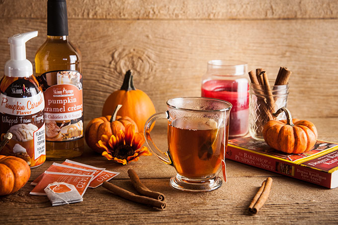 A cup of tea steeping with cinnamon stick s, pumpkins, and florals and pumpkin spice syrup on wood