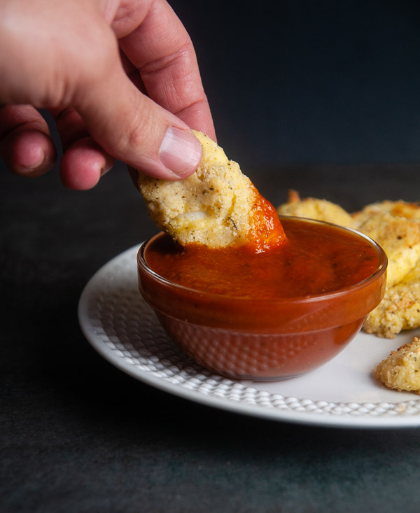 Hand dipping a low carb mozzarella stick into a bowl of sauce on a black background