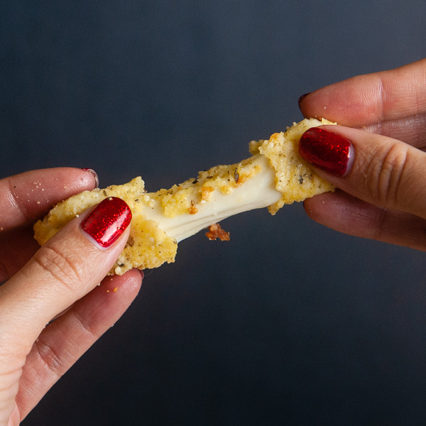Low carb mozzarella stick being stretched apart by two female hands, showing a gooey cheese pull on black