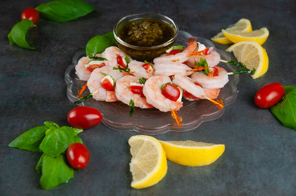 A plate full of shrimp caprese bites on a dark background with lemon slices, basil leaves, and extra cherry tomatoes