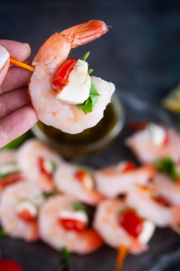 A shrimp caprese bite. A hand holding a skewer with a shrimp nestling a piece of cherry tomato and fresh mozzarella and basil on dark background above a plate of shrimp