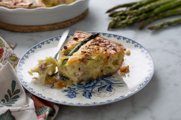 Slice of ham and asparagus quiche with fork on blue and white floral plate