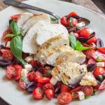 Easy Keto Greek Chicken: grilled and sliced chicken breast over a tomato salad including feta cheese, olives and fresh herbs on a platter with serving spoon.