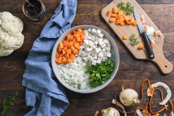 Keto shepherd's pie ingredients from left to right: cauliflower, beef base, bowl of diced carrot, turnip, onion and parsley. Cutting board with cut vegetables and knife.