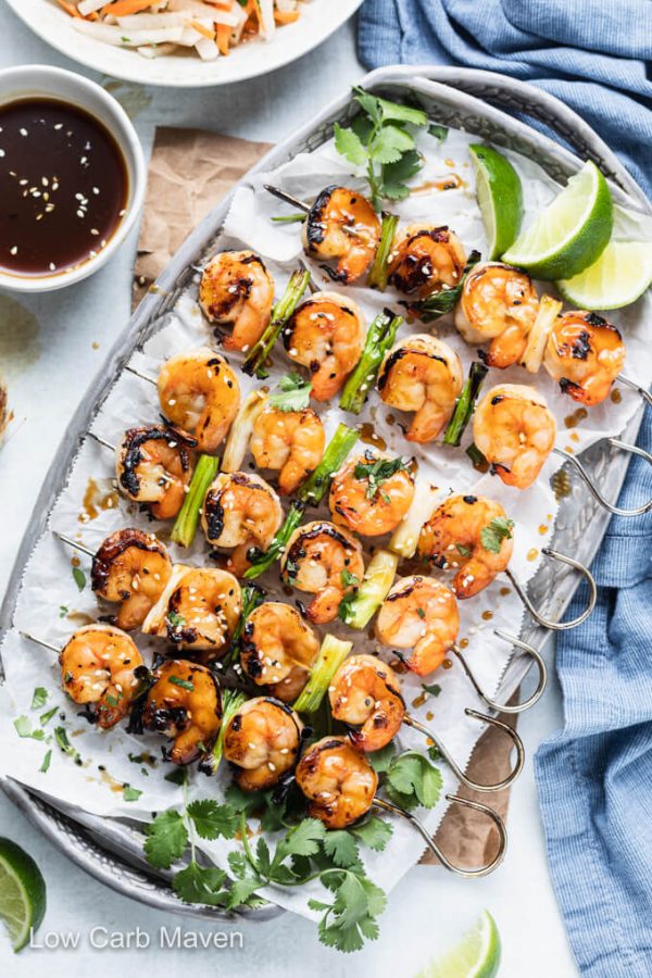 Skewered shrimp teriyaki on platter with lime and cilantro; blue napkin and bowl of teriyaki sauce.