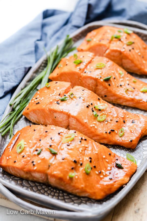 Keto salmon teriyaki garnished with sesame seeds and sliced scallions on gray platter; whole scallions and blue napkin to the left.